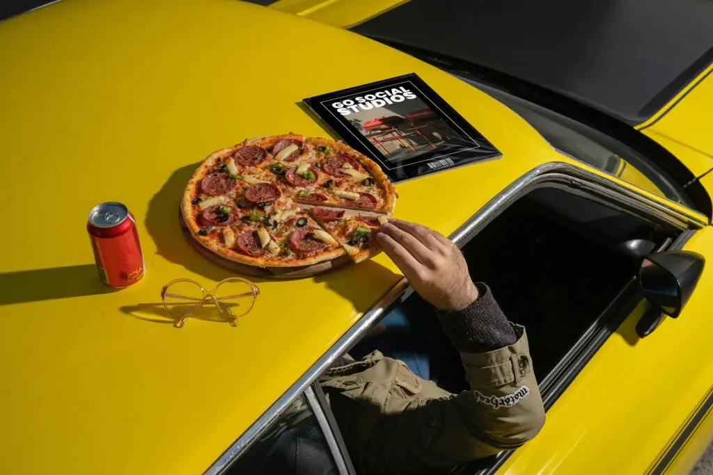 Pizza, soda, and magazine on a yellow car hood — visual metaphor for unconventional creative services offered by Go Social Studios.